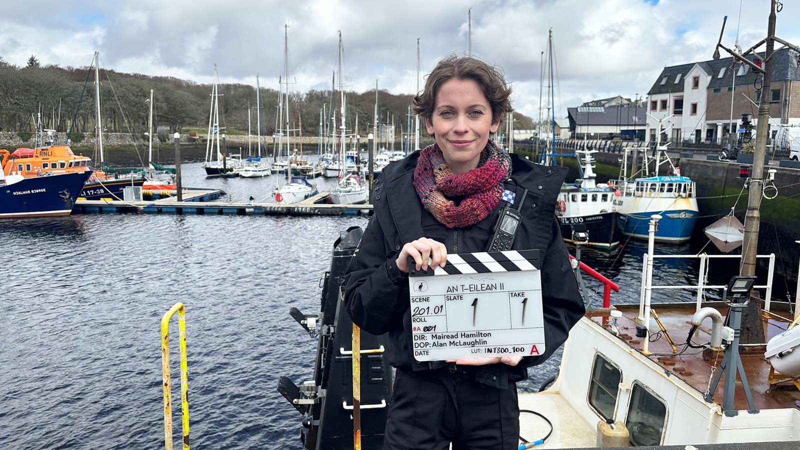 A woman stands by a harbour holding a clapperboard for "An T-Eilean II" with boats moored in the background. The sky is cloudy, and buildings line the water's edge. They wear a dark jacket and a colourful scarf.