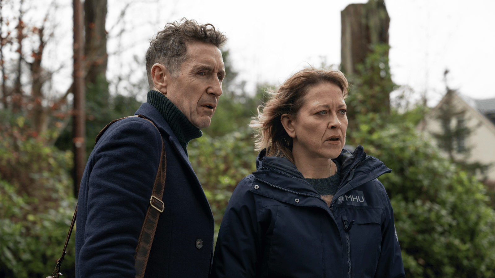 Still from Annika S2. Nicola Walker as Annika and man look concerned standing outside in front of bushes.