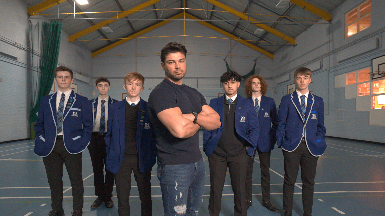 Anton Danyluk on Body Shame promotional image. Anton Danyluk stands in gym hall with school pupils standing behind him. Credit_Firecrest Films and BBC Scotland