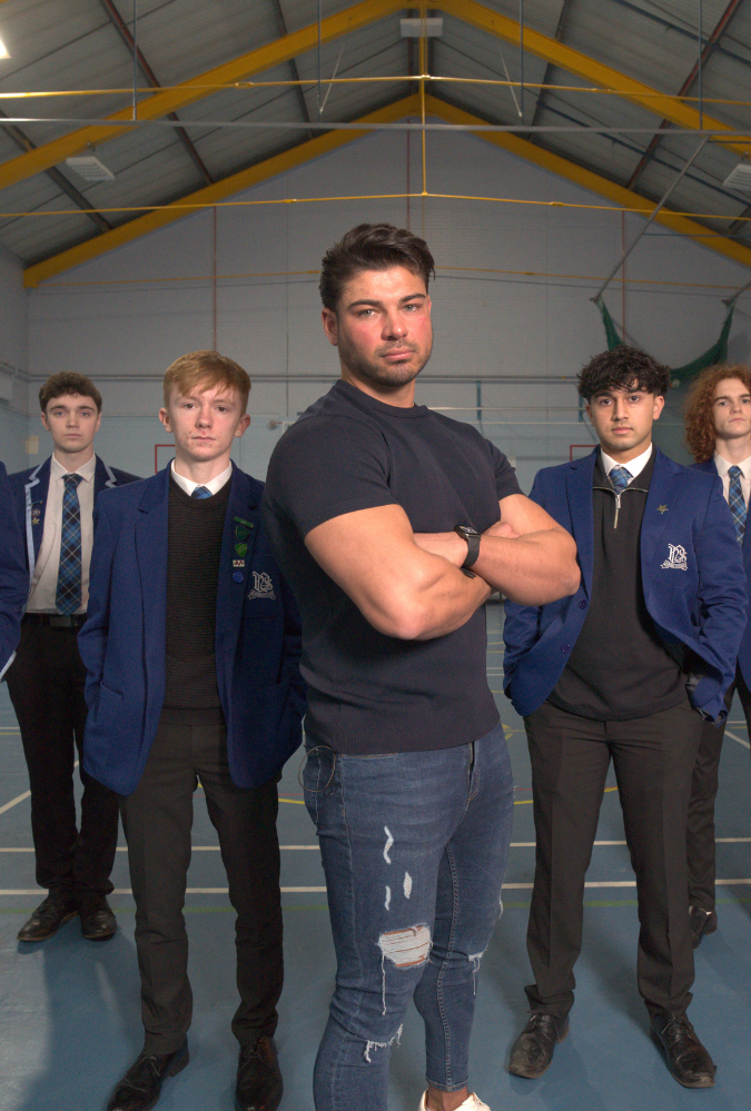 Anton Danyluk on Body Shame promotional image. Anton Danyluk stands in gym hall with school pupils standing behind him. Credit_Firecrest Films and BBC Scotland