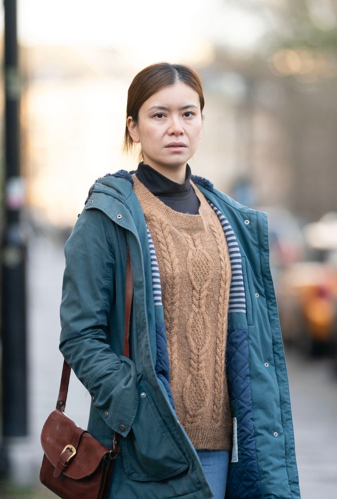 A woman stands outdoors on a street, wearing a blue coat over a brown cable-knit sweater and carrying a brown shoulder bag. The background is softly blurred with hints of buildings and trees.