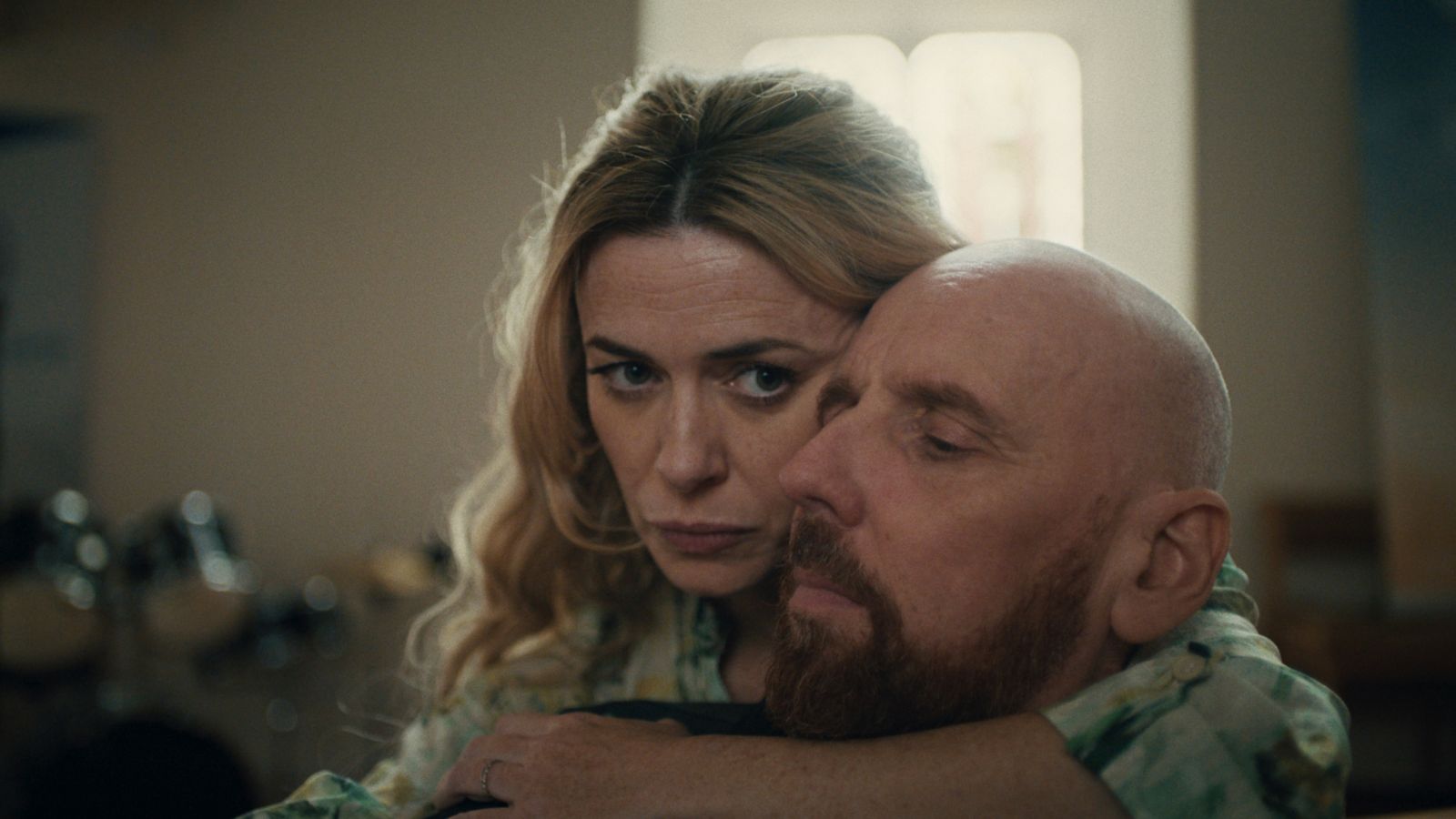 A woman with long, wavy blonde hair embraces a bald man with a beard, resting her chin on his shoulder. Her expression is serious and contemplative, while the man seems calm with his eyes closed. They are in an indoor setting with soft lighting.