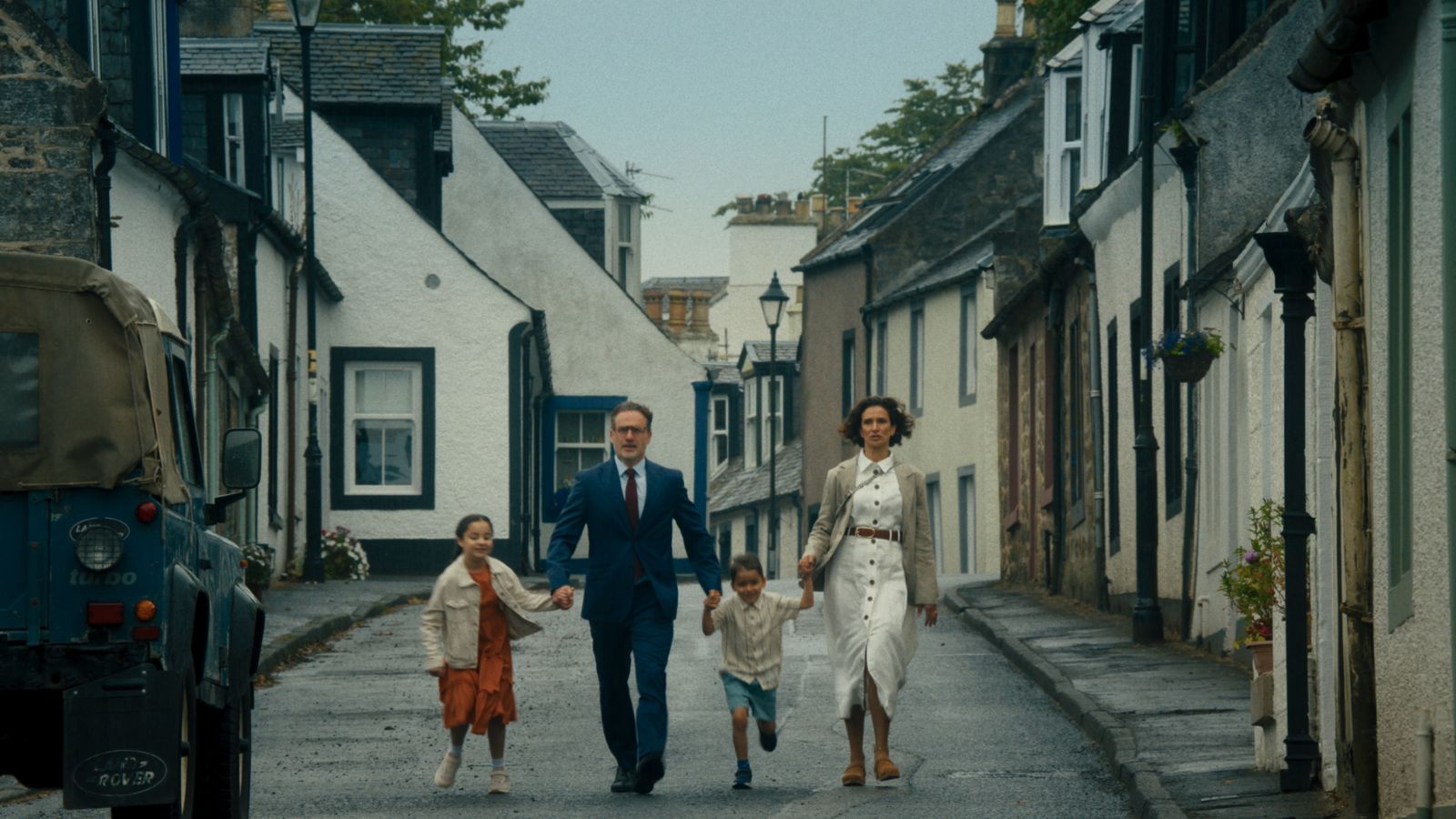 A family of four walks down a narrow street lined with quaint, stone houses. The father, in a suit, and the mother, in a white dress with a beige jacket, hold hands with their two children, a girl in a red dress and a boy in shorts. A vintage Land Rover is parked to the side.
