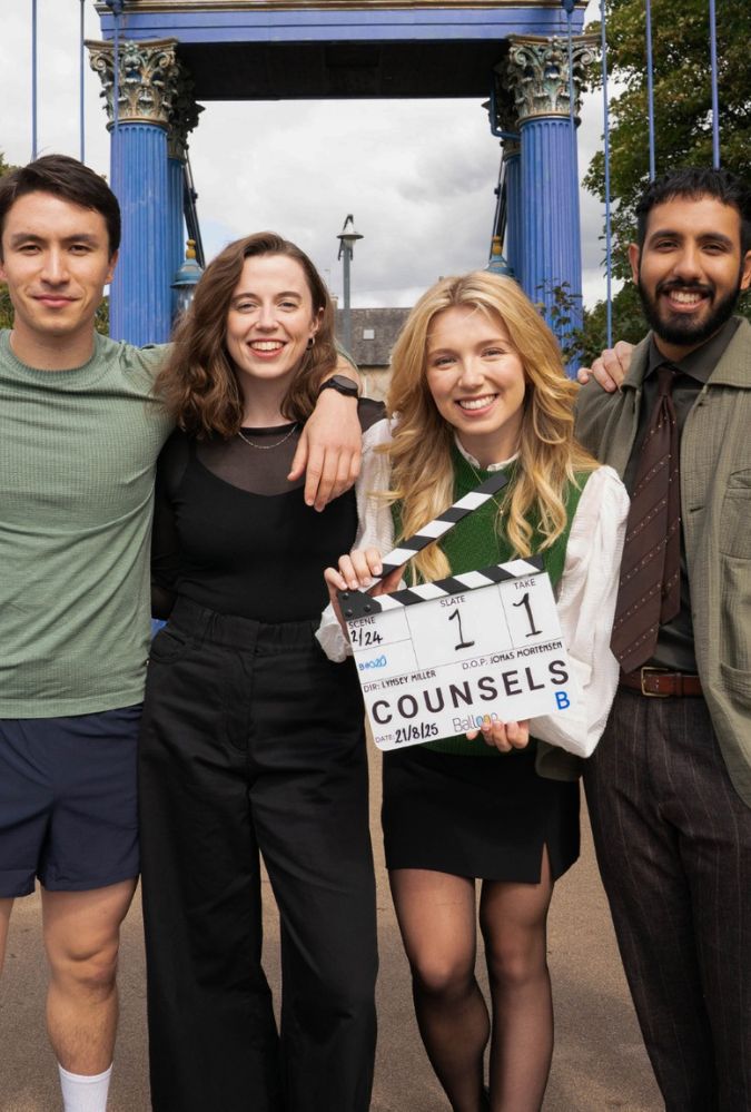 A group of four people stands together, smiling on a bridge. A young woman in the centre holds a clapperboard labeled 