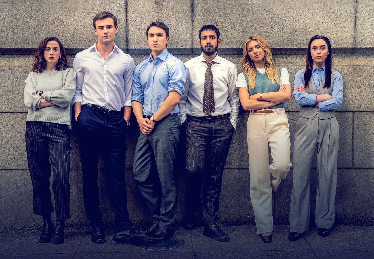 An eye-level shot captures six young professionals standing shoulder-to-shoulder, looking directly forward with serious expressions against a grey stone block wall.