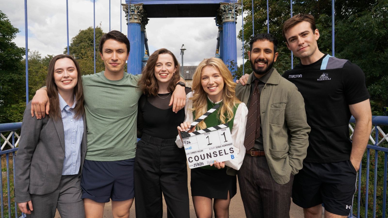 A group of six people stands together, smiling on a bridge. A young woman in the centre holds a clapperboard labeled "Counsels" with scene details. Behind them, the blue structure of the bridge is visible against a backdrop of trees and a cloudy sky. They appear cheerful and casually dressed.