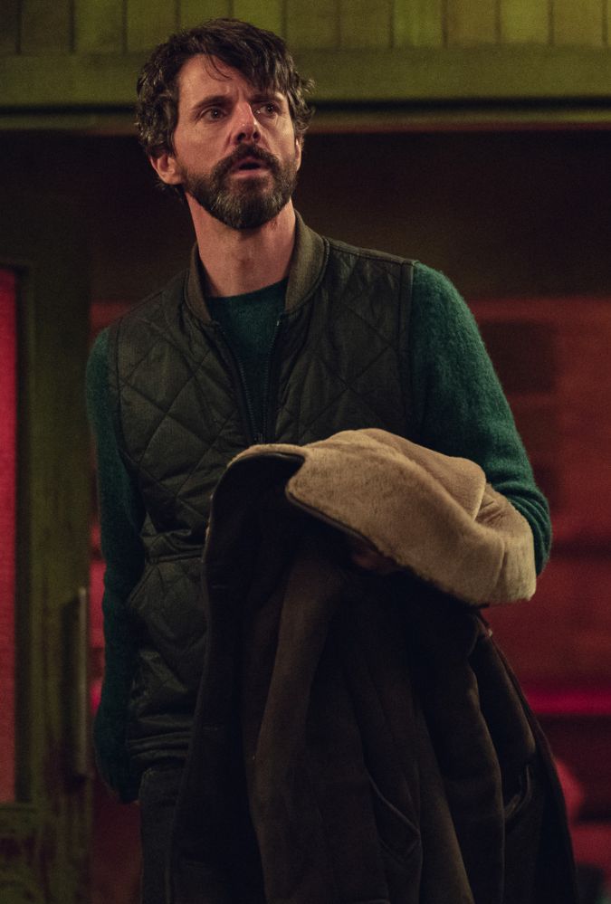 A person stands in a dimly lit room, holding a coat, surrounded by green walls and a red-lit doorway.