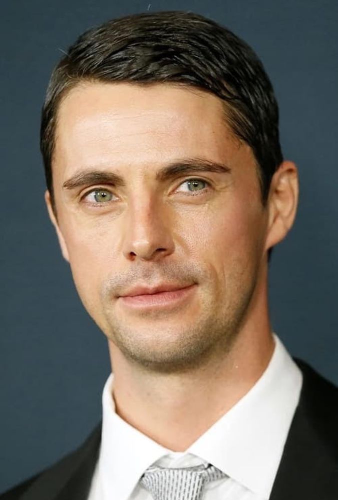 A headshot of actor Matthew Goode from the shoulders up. He wears a black suit, white shirt and a silver tie. Courtesy of Netflix