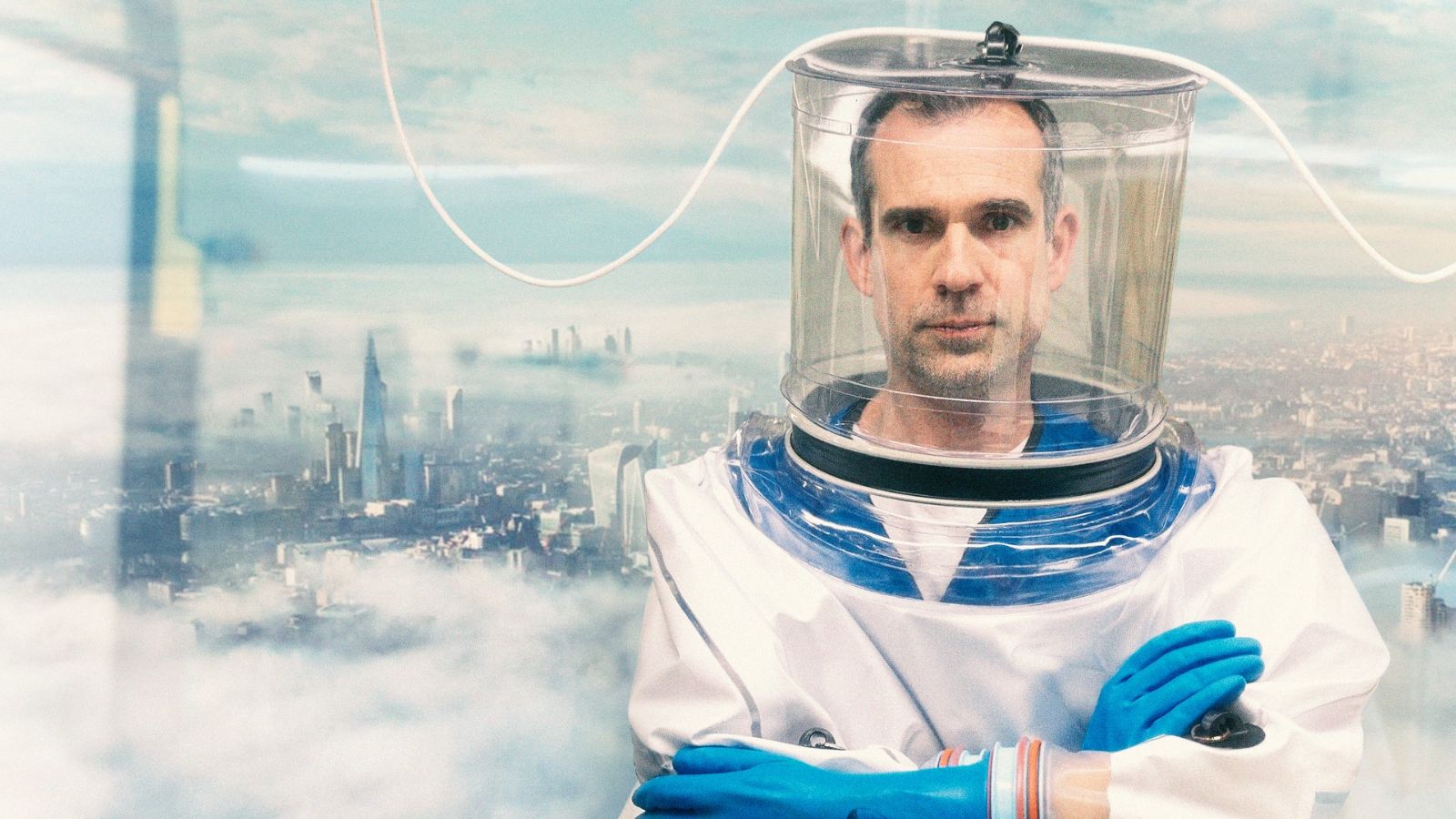 A person wearing a white hazmat suit with a transparent helmet and blue gloves stands against a cityscape backdrop. The skyline, including the Shard, is visible through a foggy atmosphere.