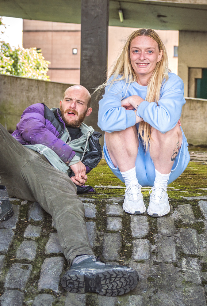 Blonde woman and tman sit together. They appear to be in a residential estate under a bridge.