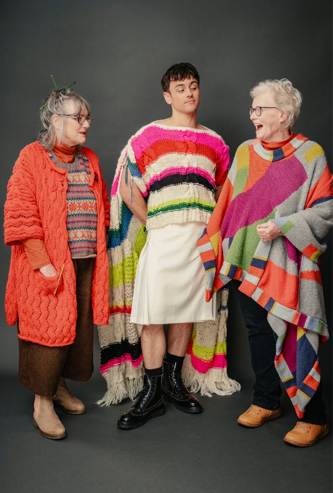 Three people stand together wearing colourful, oversized knit garments. The middle person wears a vibrant striped poncho and a white skirt, flanked by two older individuals in multicoloured sweaters. The left person is in an orange cardigan, and the right in a gray and pink poncho. All are smiling, set against a dark background.