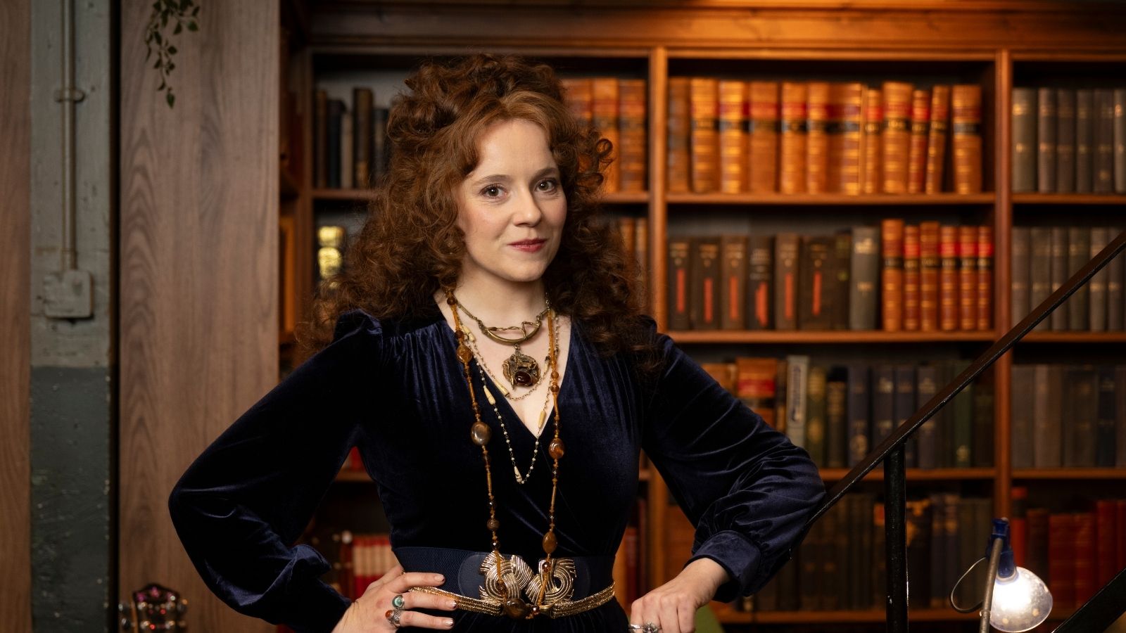 A woman with curly red hair stands confidently with hands on hips, wearing a dark blue velvet dress adorned with ornate jewellery. She is in front of shelves filled with neatly arranged books, creating a warm, intellectual backdrop.