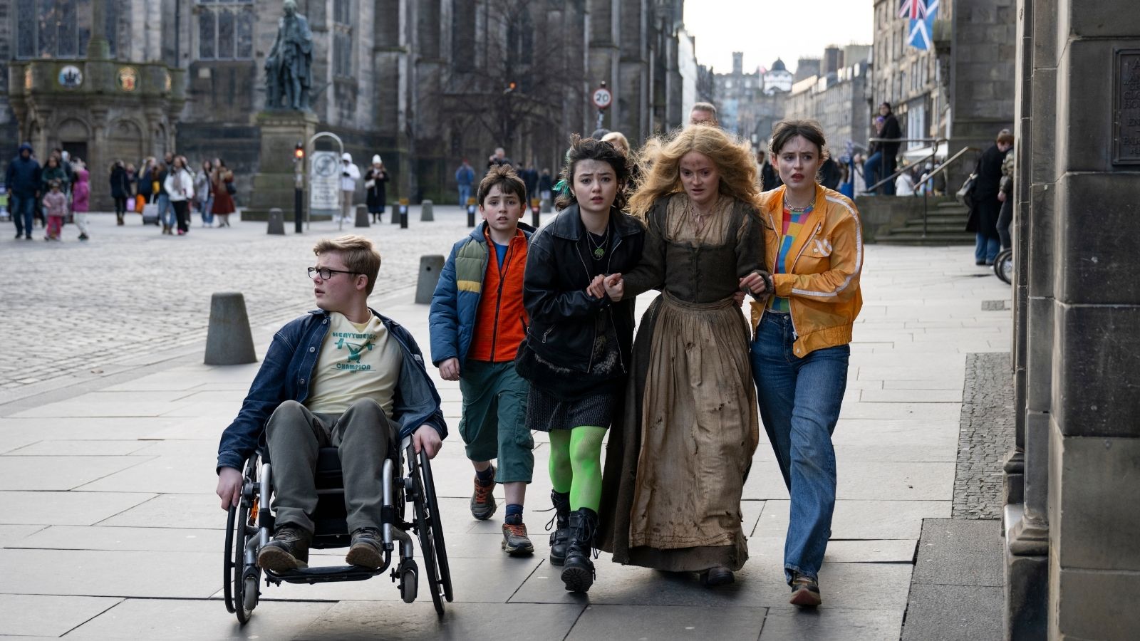 A group of five individuals walk down a street. One person is in a wheelchair, while others surround a woman in a vintage dress, guiding her. The background features a historic stone building and a statue, with other pedestrians visible. Flags are seen in the distance.