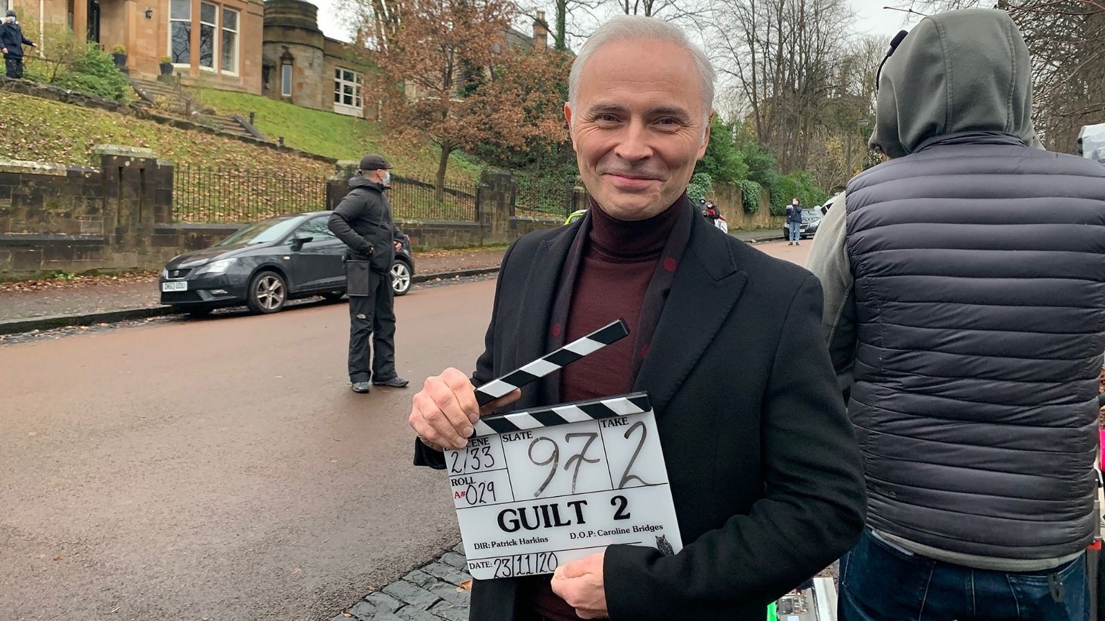 Mark Bonnar, star of Guilt, holding a clapper board and smiling on a street.