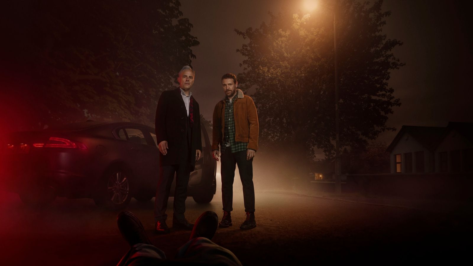 Mark Bonnar and Jamie Sives stand on a suburban road in Guilt, with a pair of feet or a body lying on the ground ust in shot