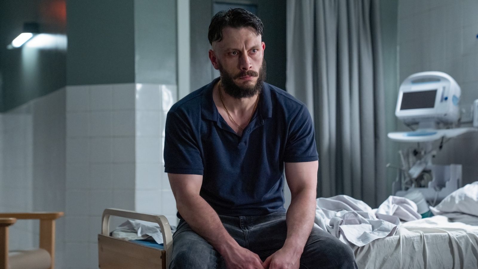 A man with a beard and short hair sits on the edge of a hospital bed, wearing a navy blue polo shirt and dark pants. He appears serious and contemplative. The room contains medical equipment, with tiled walls and a curtain in the background.