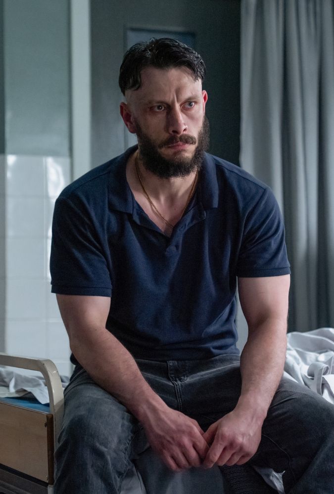 A man with a beard and short hair sits on the edge of a hospital bed, wearing a navy blue polo shirt and dark pants. He appears serious and contemplative. The room contains medical equipment, with tiled walls and a curtain in the background.