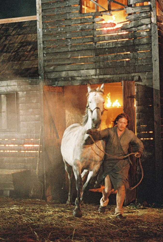 Still from Harvest, credit Jaclyn Martinez. Actor Caleb Landry-Jones leads a white horse by the reigns away from a burning wooden building at night