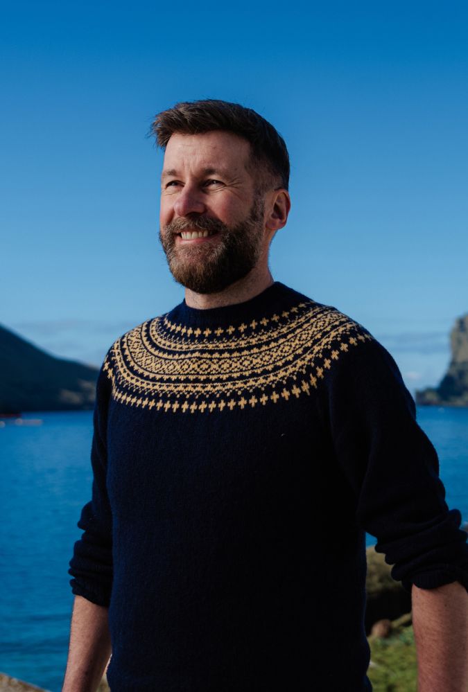 A man with a beard, wearing a navy-blue sweater with intricate patterns and a kilt, stands smiling in front of a scenic coastal landscape.
