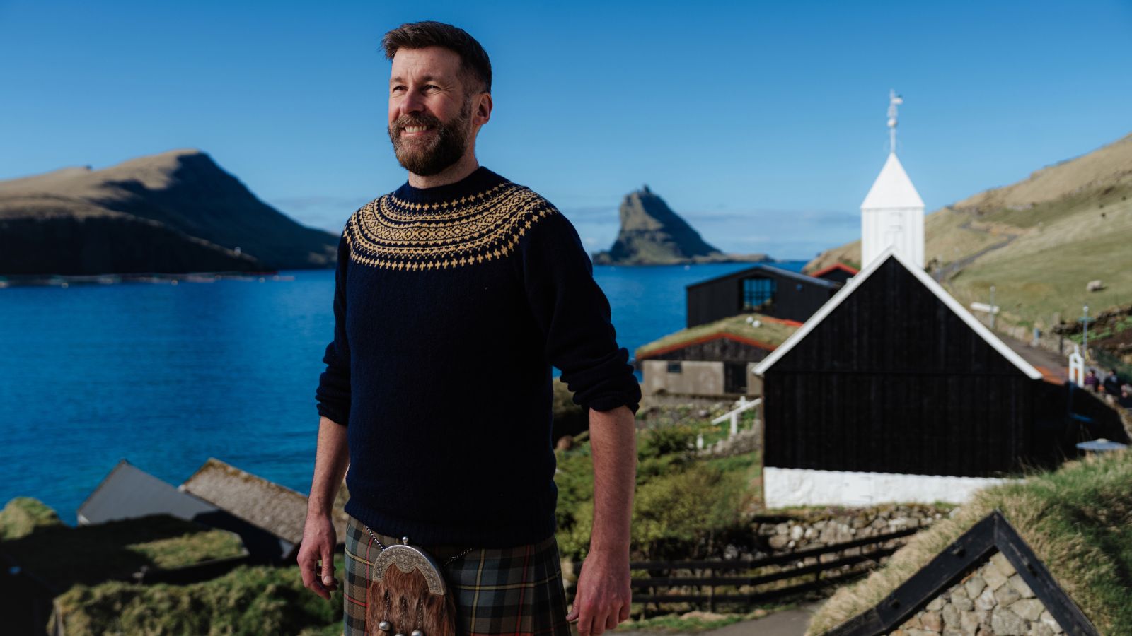 A man with a beard, wearing a navy-blue sweater with intricate patterns and a kilt, stands smiling in front of a scenic coastal landscape. There's a calm blue sea, grassy hills, and a small white church with a pointed steeple in the background under a clear blue sky.