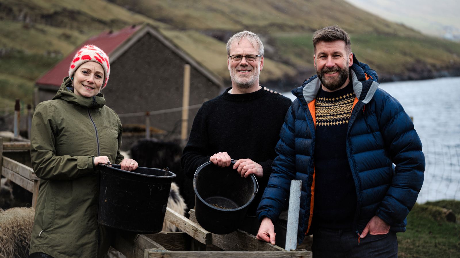 A group of three people stand outdoors beside a wooden fence, holding black buckets. The woman on the left wears a green jacket and a red-patterned beanie. The man in the middle has short gray hair, glasses, and a black sweater. The man on the right wears a blue puffer jacket with a patterned sweater underneath. The background features grassy hills and a body of water.
