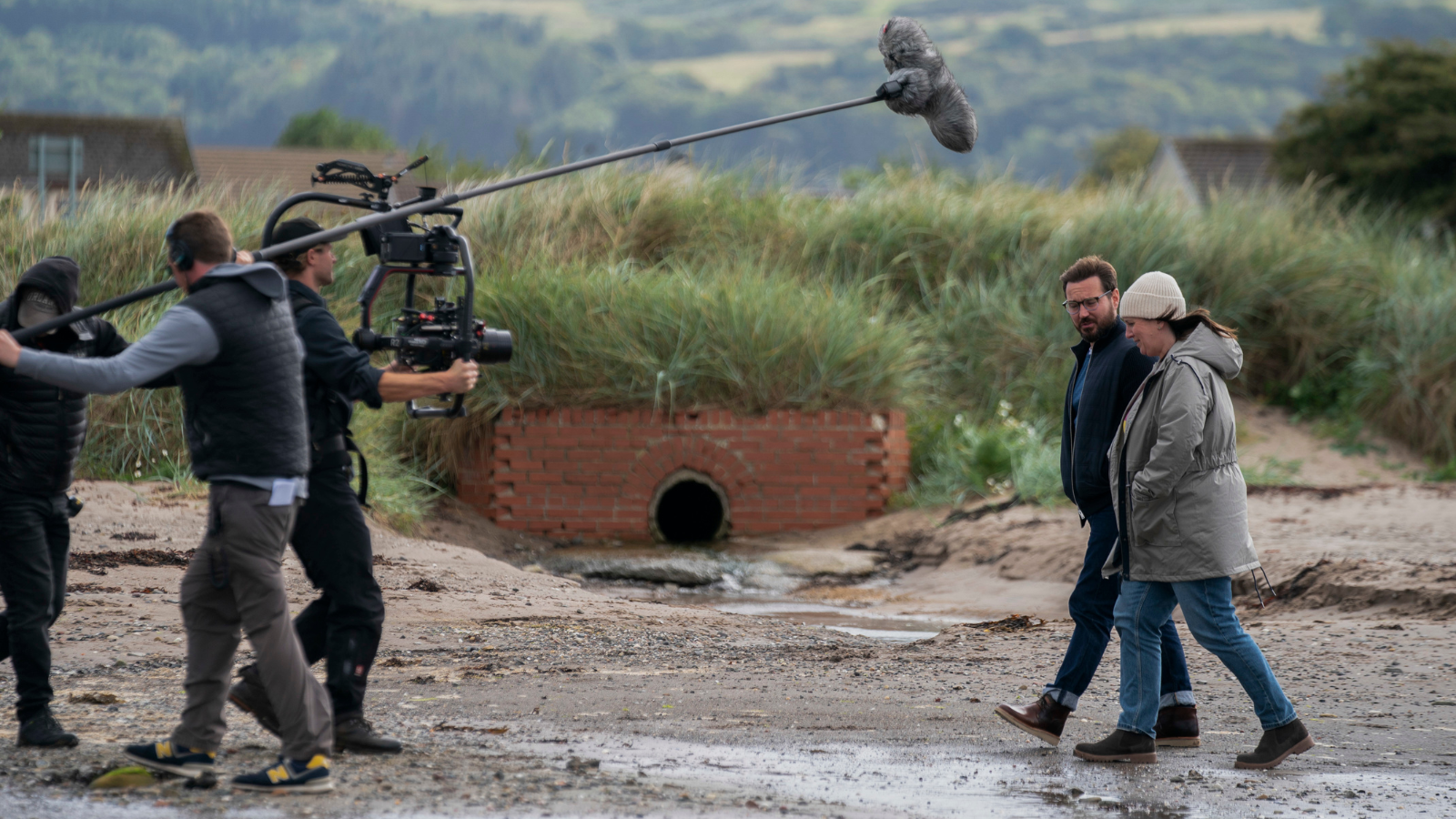 Behind the scenes image from Mayflies. Man and woman walk a long a wet beach while film crew shoot them from the front.