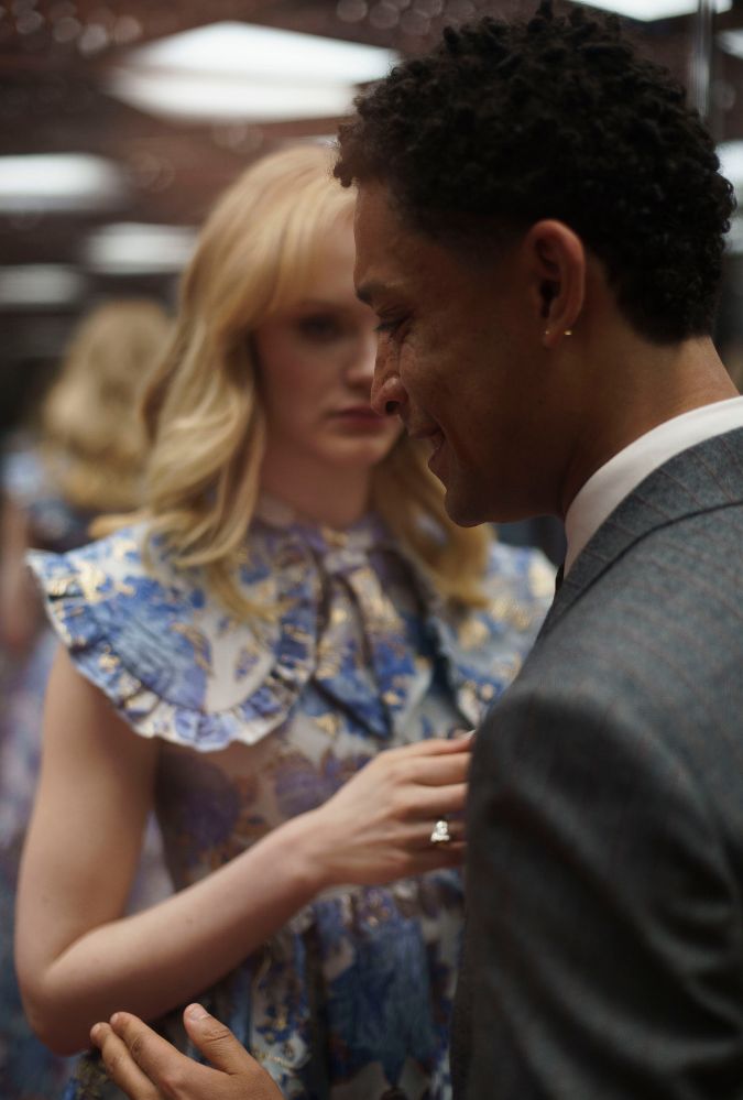 A woman in a floral dress with ruffled sleeves stands facing a man in a suit. The scene is intimate and softly lit, with the woman's hands gently touching the man's chest. Their expressions are focused, creating a moment of quiet connection.