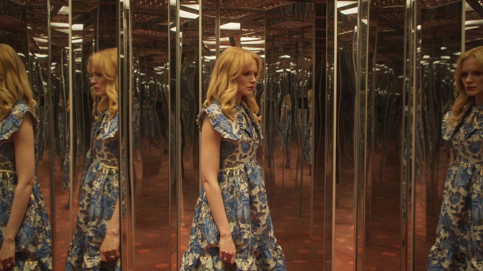 A woman in a floral dress stands in a room full of mirrors, her reflection repeated countless times. The room has a warm tone, and the floor has a subtle pattern. Her expression is contemplative, and the light casts soft shadows.