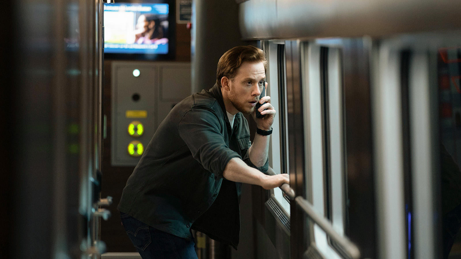 A man in a green shirt with rolled up sleeves talk on the phone as he looks out of a train window