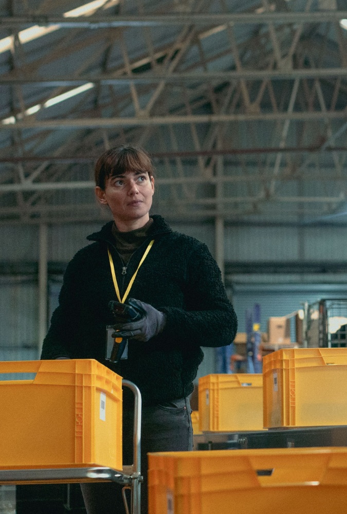 Still from On Falling, courtesy of Sixteen Films. A woman, played by actress Joana Santos stands in a large warehouse, with yellow crates stacked on tables all around her.