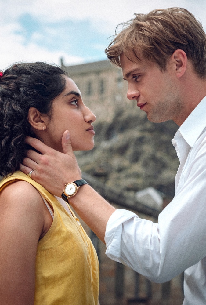 The still from Netflix production One Day, features a man, Dexter played by actor Leo Woodall and a woman, Emma, played by actress Ambika Mod, looking at each other, with Dexter's arm caressing Emma's neck and chin. Behind them is part of Edinburgh Castle.