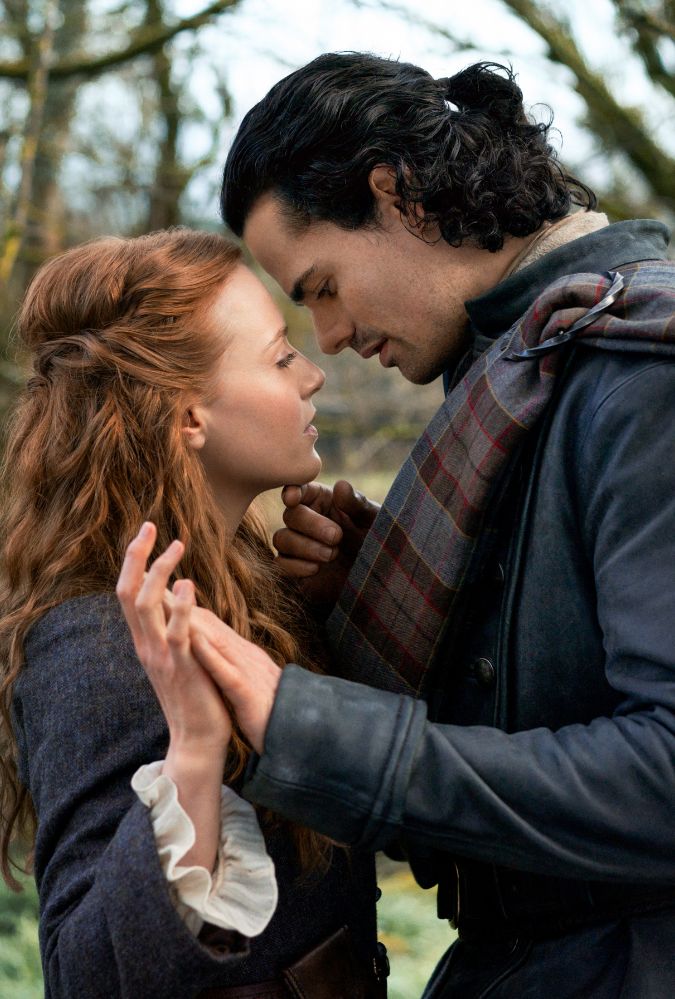 A man and woman stand closely, gently holding hands while gazing into each other's eyes. The man has curly dark hair and wears a tartan shawl over a coat. The woman has long, wavy red hair and wears a dark blue coat with white ruffled cuffs. The background features blurred trees, suggesting an outdoor setting.