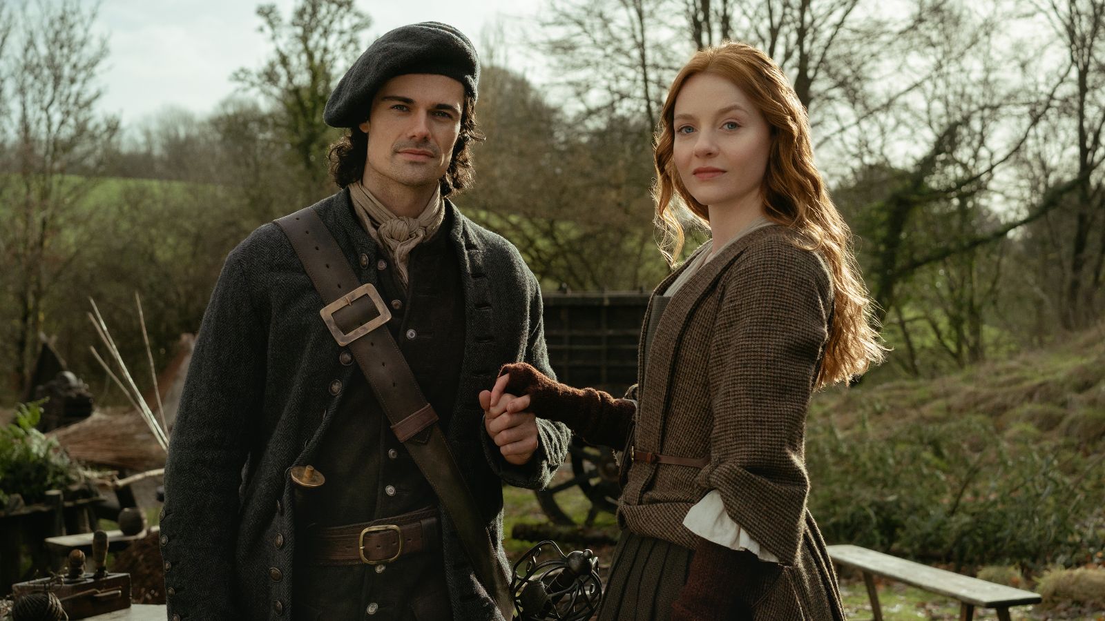 A man and woman stand outdoors in historical clothing, against a backdrop of trees and rustic items. The man wears a dark beret and coat with a leather strap, while the woman wears a long brown dress and gloves. They hold hands, gazing confidently at the camera. The scene has a vintage, pastoral feel.