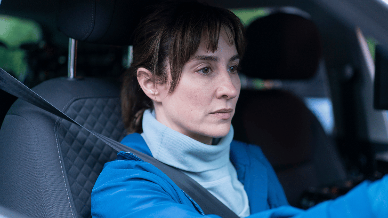 Morven Christie in Payback. Middle aged women with dark hair tied in a pony tail driving a car.