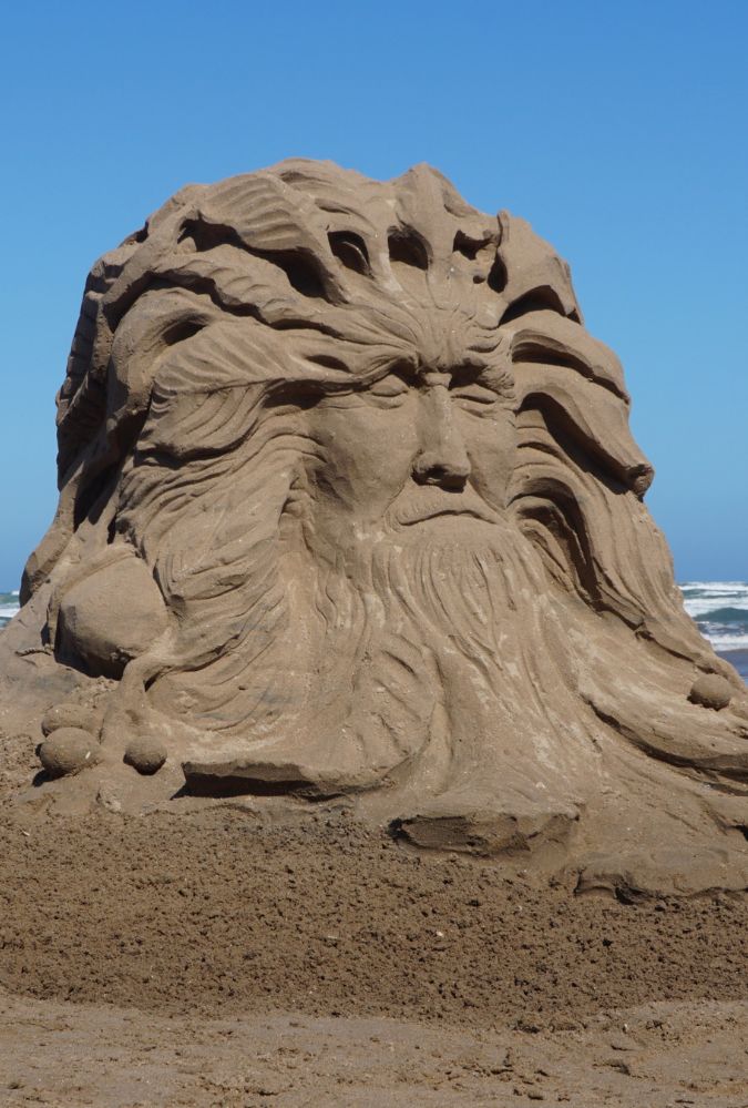 An outdoor shot captures a large, detailed sand sculpture of an older man's face on a beach, under a clear blue sky.