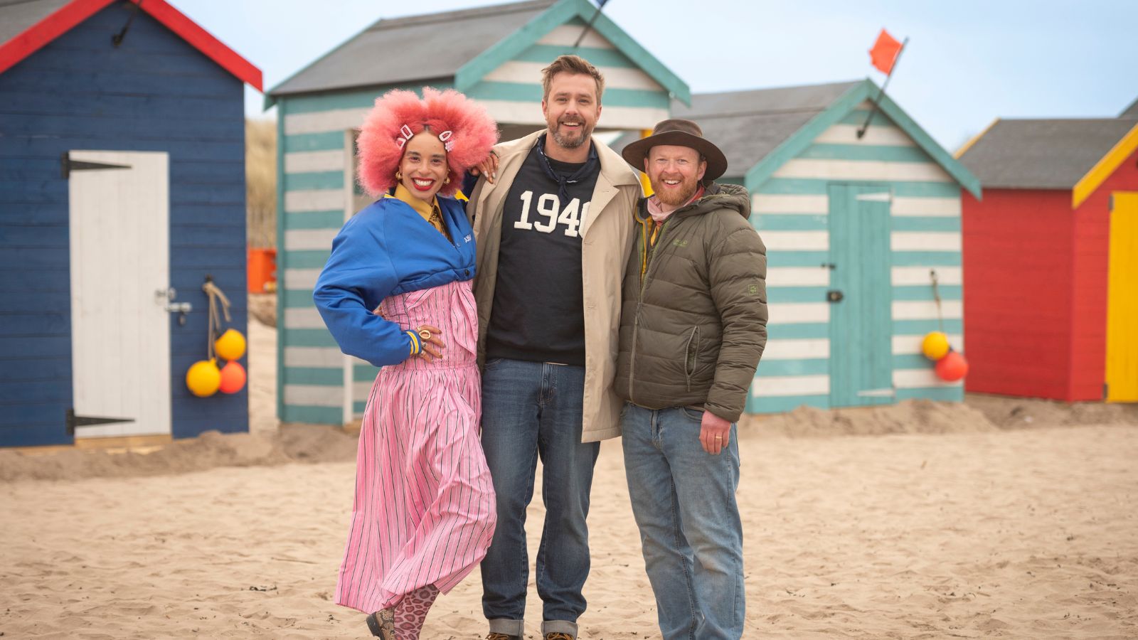 An outdoor medium shot shows three smiling people standing arm-in-arm on a sandy beach in front of a row of colourful beach huts.