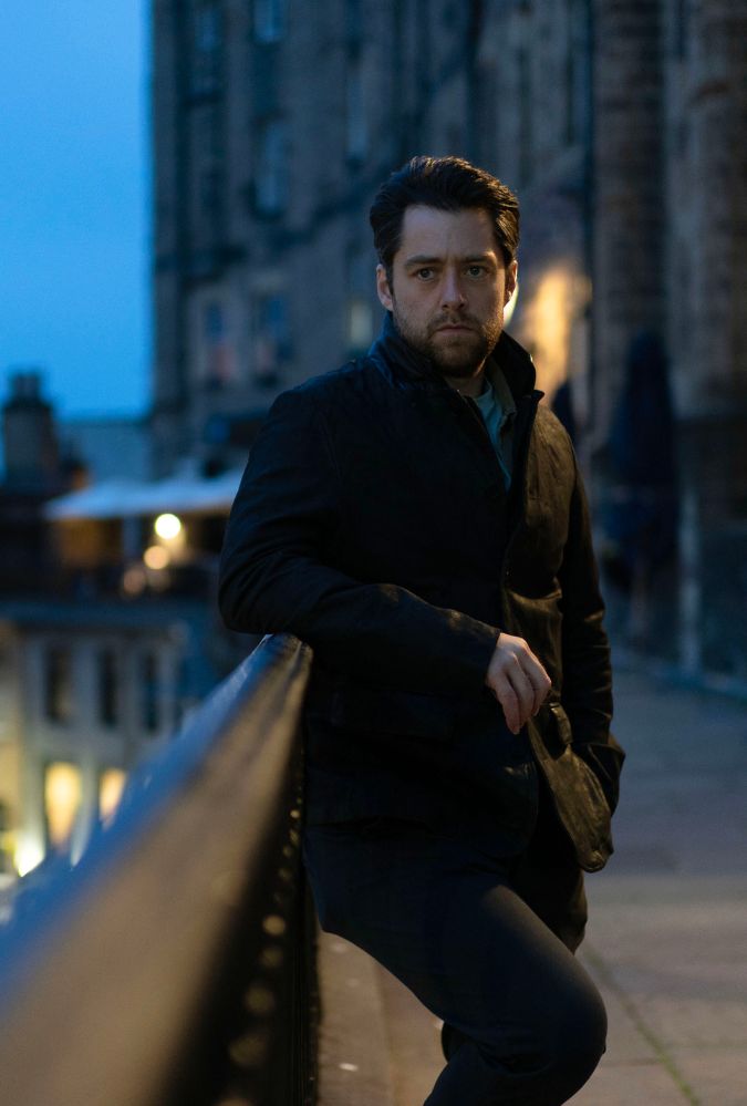 till from Rebus, courtesy of BBC. Richard Rankin wears black jacket and trousers and leans on a railing on Victoria Terrace in Edinburgh.
