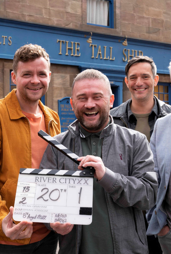 Cast of River City's 20th Episode celebrations standing on set, holding a clapper board.