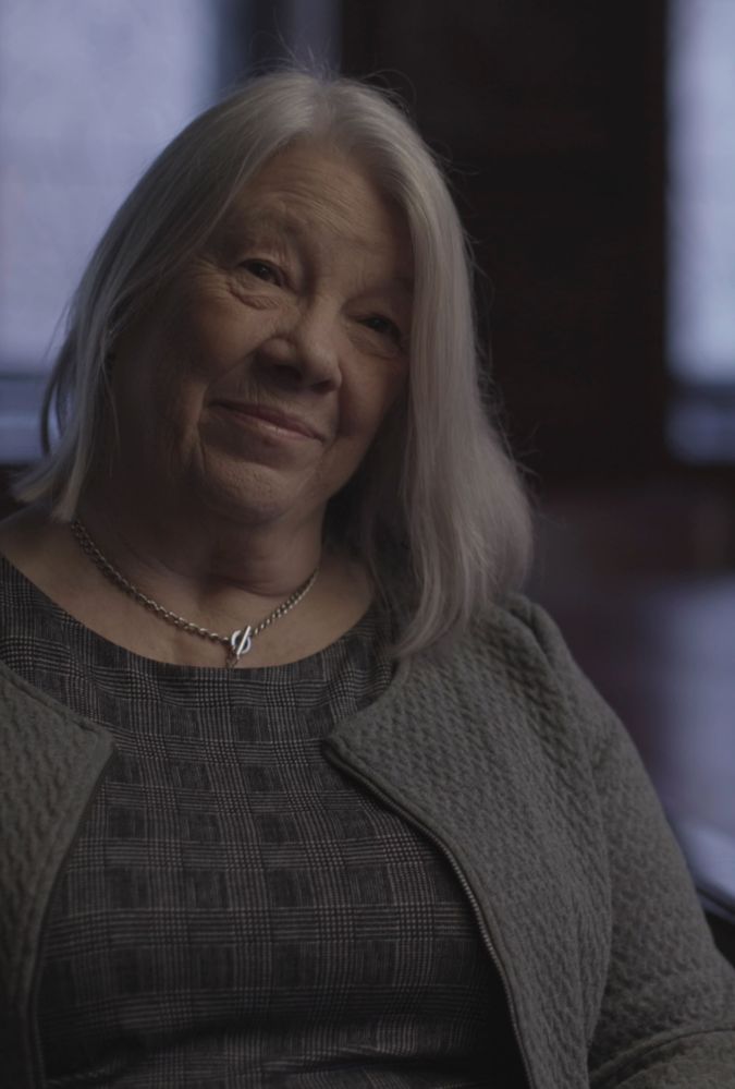 An medium close-up shot of a white-haired woman with a gentle smile sitting in a dimly lit, wood-panelled room. She is positioned on the left side of the frame, looking slightly off-camera with a warm, thoughtful expression. She wears a grey patterned top beneath a textured, open-front grey jacket and a simple silver necklace. The background features a long, polished wooden table stretching to the right, with blurred windows and chairs in the soft, dark background.