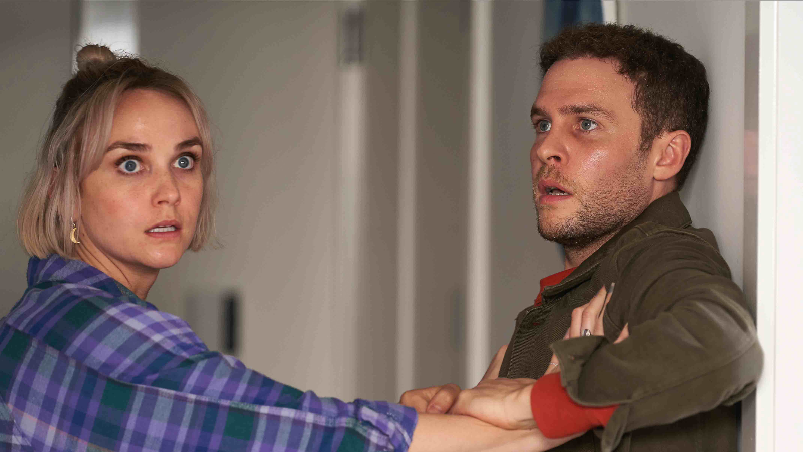 Man and women look distressed. Woman with blonde hair grabs man's coat as he stands up against a wall.