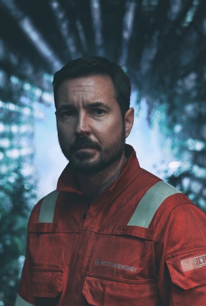 Mark Compston plays a rig worker, wearing an orange boilersuit, looking seriously at the camera with a light shining from behind him