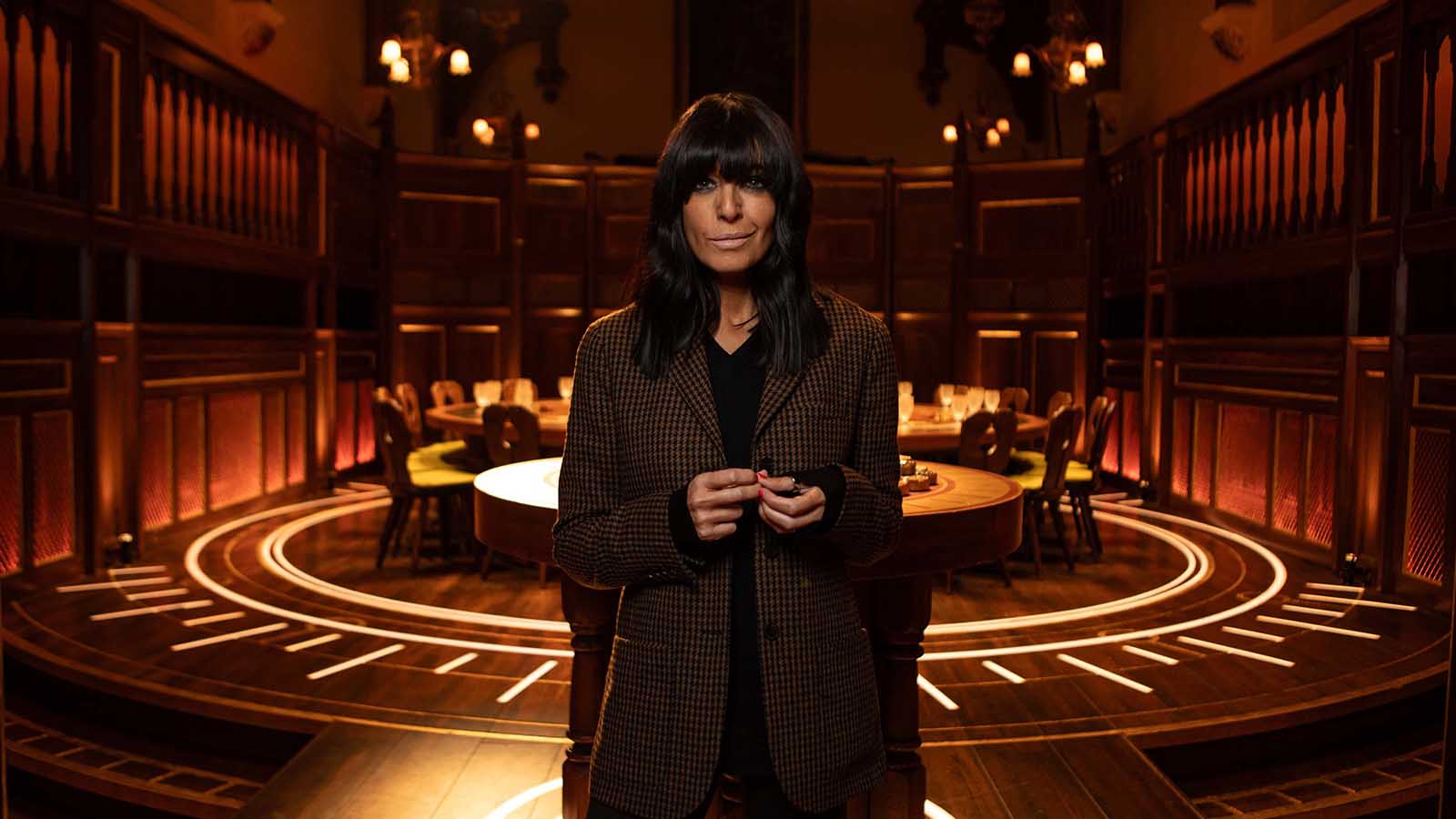 Presenter Claudia Winkleman stands in a grand room, lit dramatically, with a large table and many chair around it