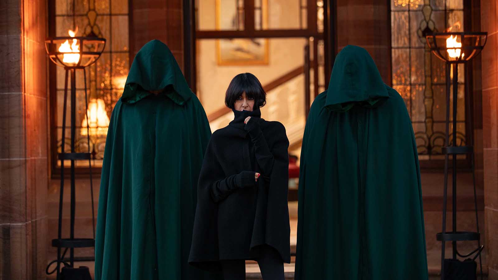 Header image for series 2 of The Traitors (courtesy of the BBC). Alt: Presenter Claudia Winkleman stands before a grand stone doorway, flanked by two tall figures on either side of her, wearing dark robes with hoods that obscure their identities