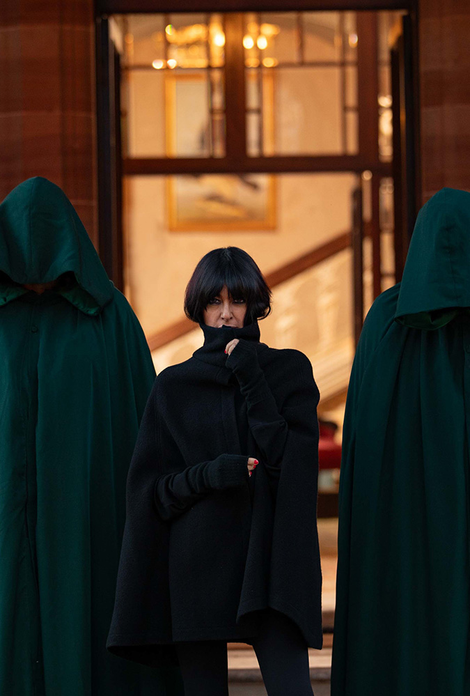 Header image for series 2 of The Traitors (courtesy of the BBC). Alt: Presenter Claudia Winkleman stands before a grand stone doorway, flanked by two tall figures on either side of her, wearing dark robes with hoods that obscure their identities