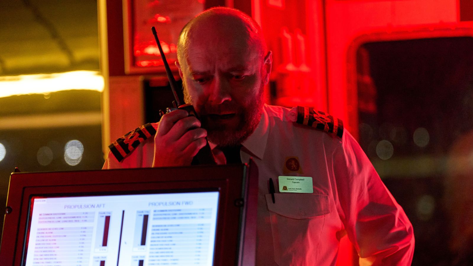 Under intense red lighting, a male ship captain with a shaved head and beard speaks urgently into a walkie-talkie. He wears a white uniform shirt with epaulets, a black tie, and a name tag that reads "Donald Campbell, Captain." In the foreground, a computer monitor displays propulsion data screens with bar graphs.