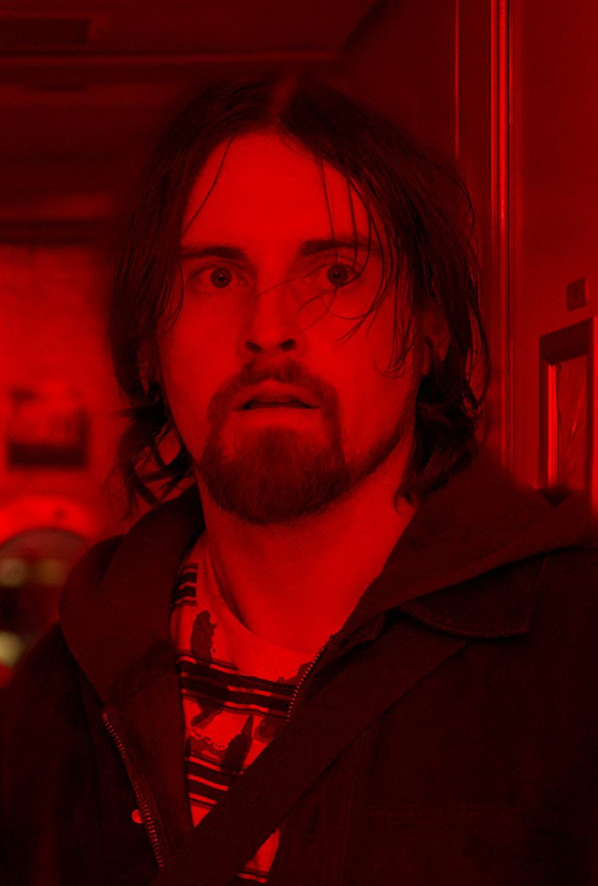 A man stands in a narrow hallway bathed in intense red light. A circular window is visible at the end of the hall.