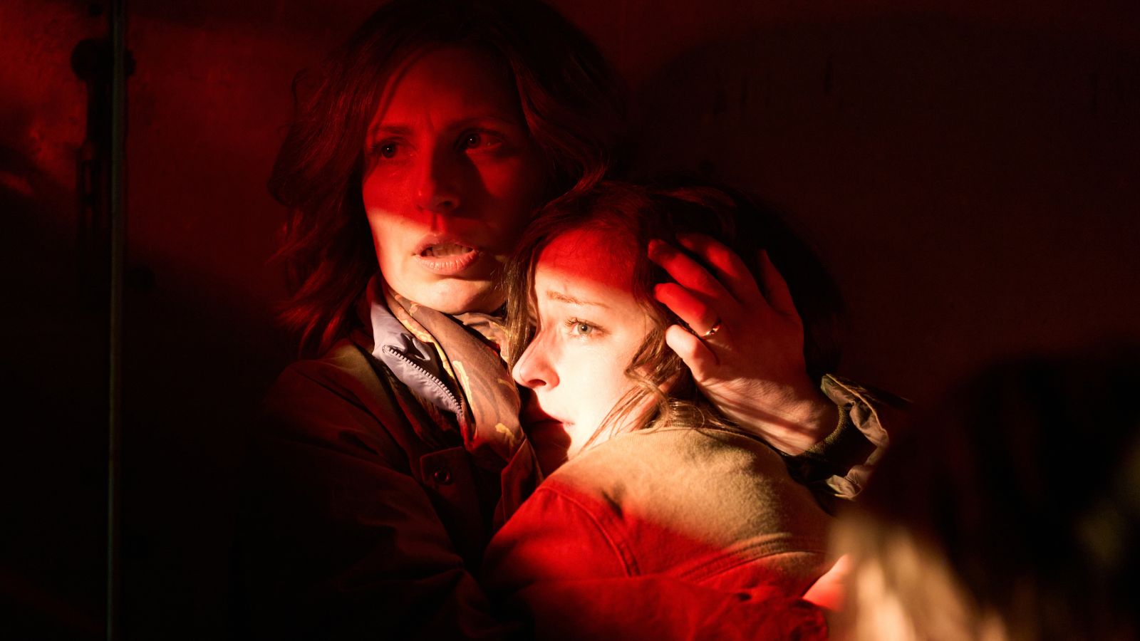 A close-up shot under dramatic red lighting shows a mother holding her daughter tightly in a protective, tense embrace.
