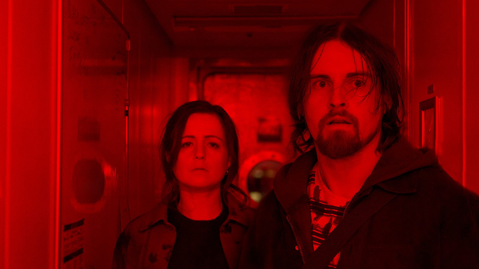 Two people stand in a narrow hallway bathed in intense red light. Both have expressionless faces, one with long hair and a beard, the other with shoulder-length hair. A circular window is visible at the end of the hall.