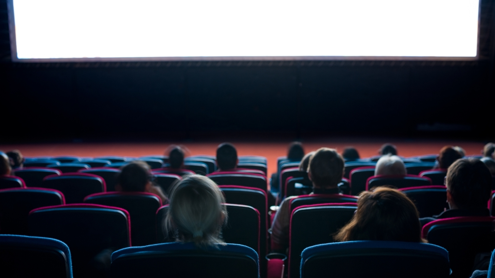 Rearview of a cinema audience section - there is a small group of people spread out on different cinema seats that are red and black. A large white cinema screen sits in front of them