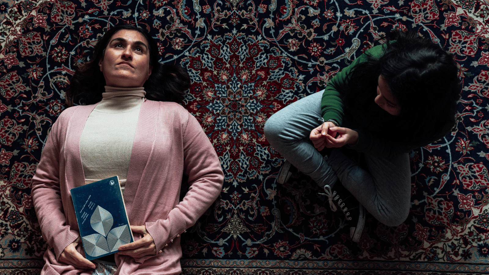 Still from BAHAAR short film. Woman in pink cardigan lies on ground holding book. Young girl sits cross legged looking at her.